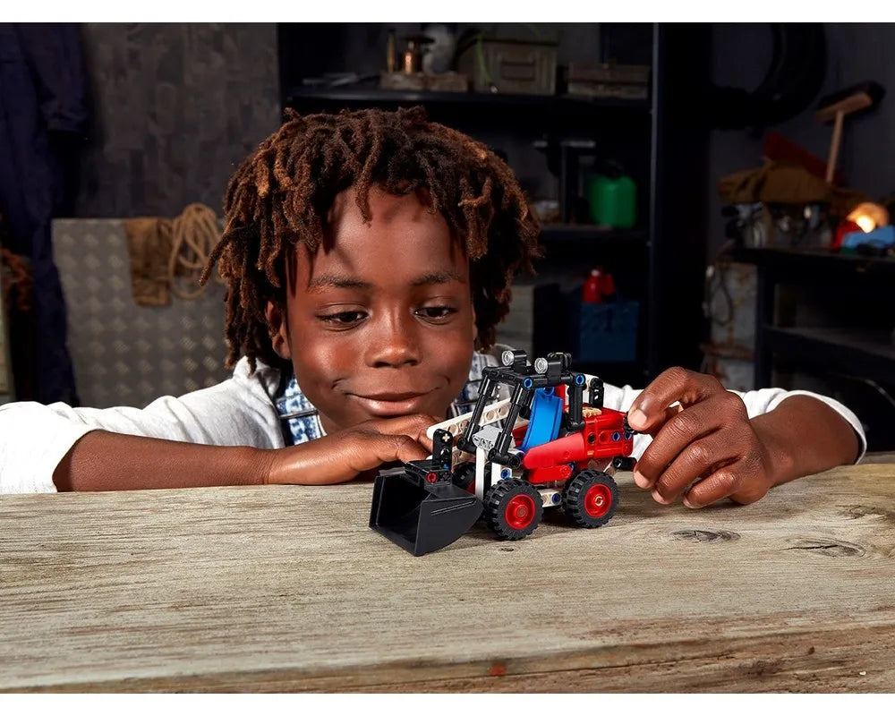 Young child admiring a red, blue, and black LEGO Technic 42116 Skid Steer Loader model on a wooden table.