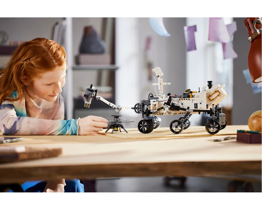 Young girl examines a LEGO Technic NASA Mars Rover Perseverance model with its Ingenuity helicopter on a wooden desk.