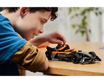 Young boy smiling and adjusting the orange and black LEGO Technic 42169 NEOM McLaren Formula E Team car model on a table.