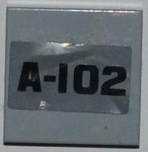 Light gray LEGO 2x2 tile with a silver sticker displaying the black text 'A-102'.