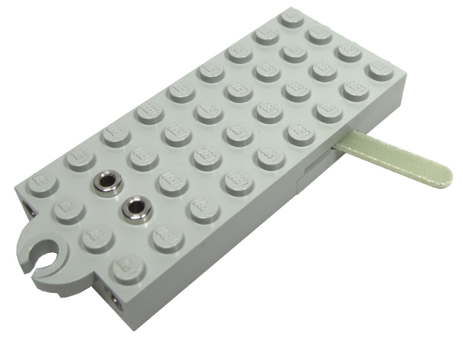 A light gray LEGO 4x9 electric train pole reverser brick with two metal rings, a magnet holder, and a green strap.